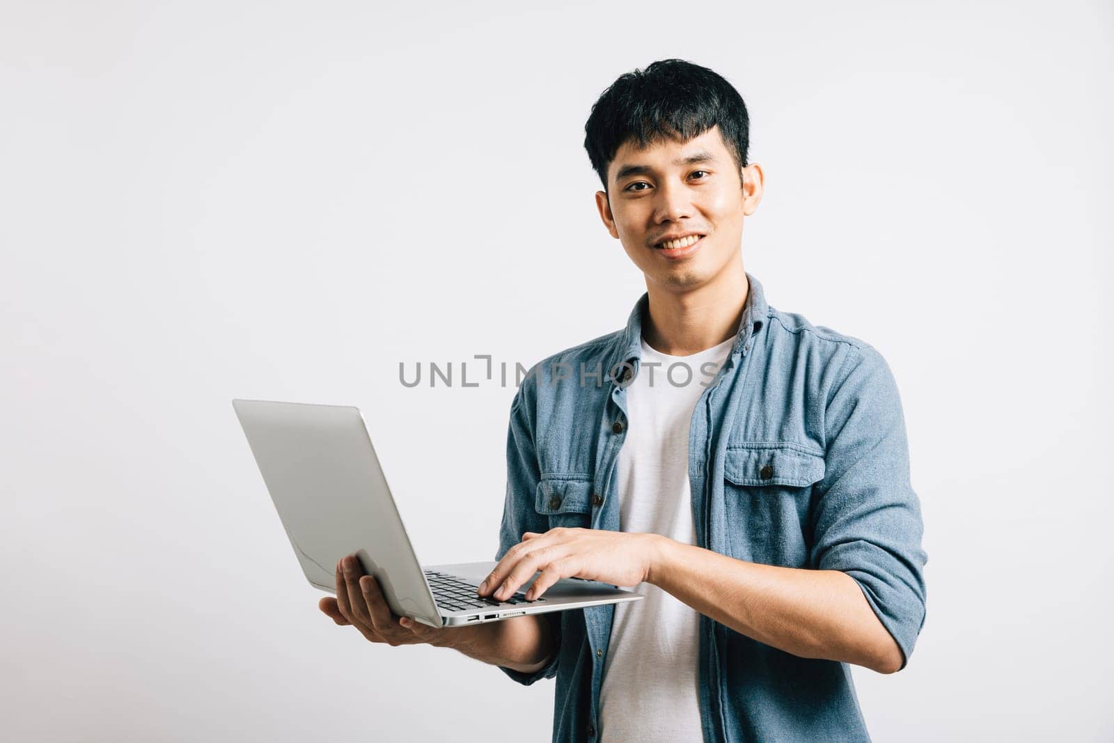 A smiling man, full of confidence, types on a laptop keyboard, engaged in successful email or chat conversations. Studio shot of an Asian businessman isolated on white, embracing technology.