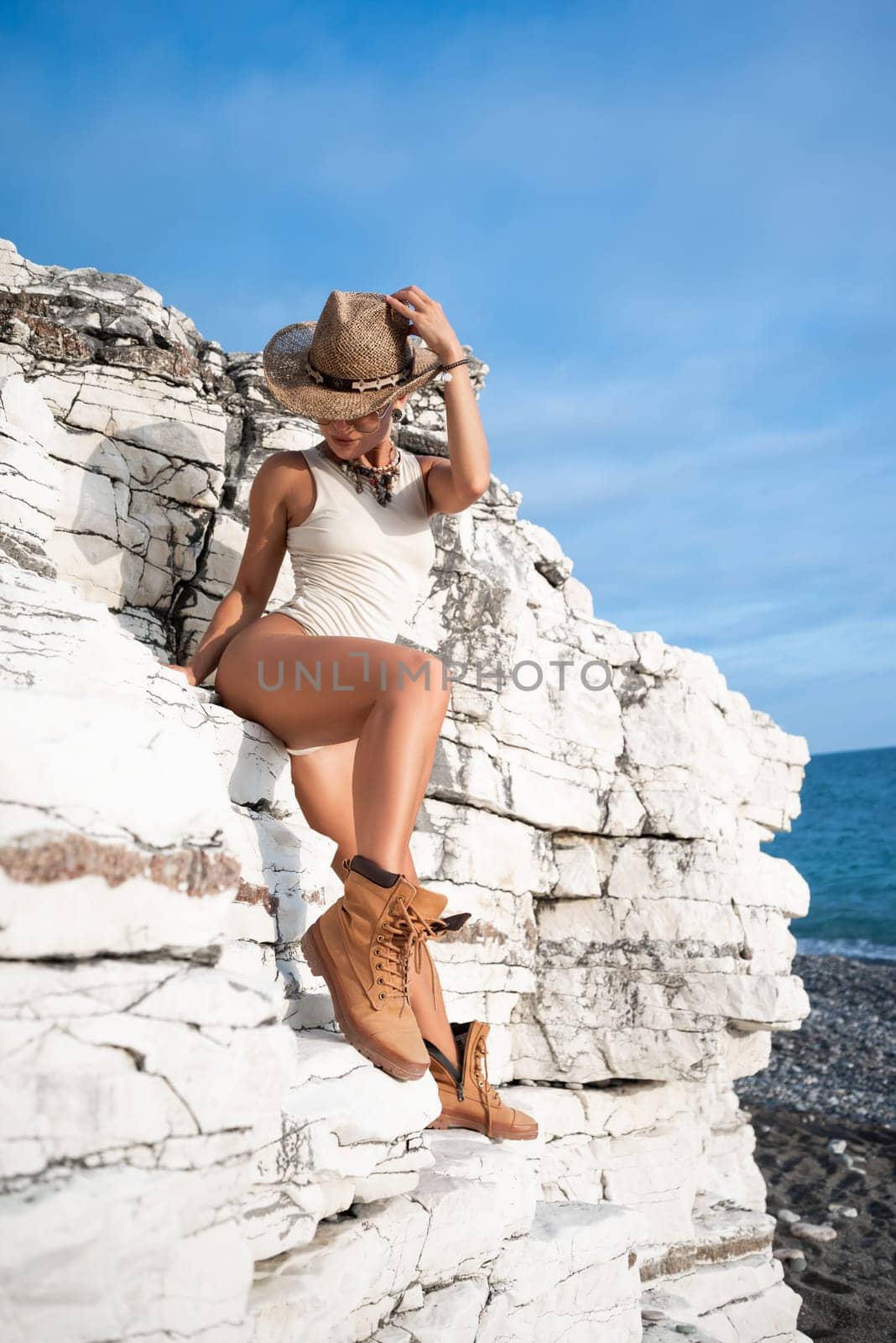 sexy girl in a bodysuit and a cowboy hat and cool boots poses freely sexually on white rocks
