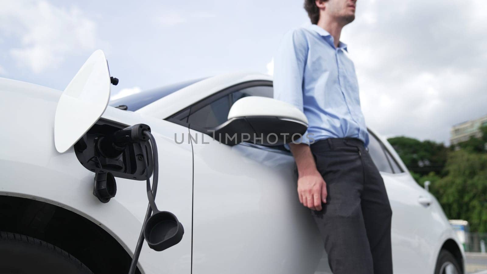 Suit-clad businessman with progressive ambition leaning on his electric vehicle while standing on a charging station with a power cable plug and a renewable energy-powered electric vehicle.