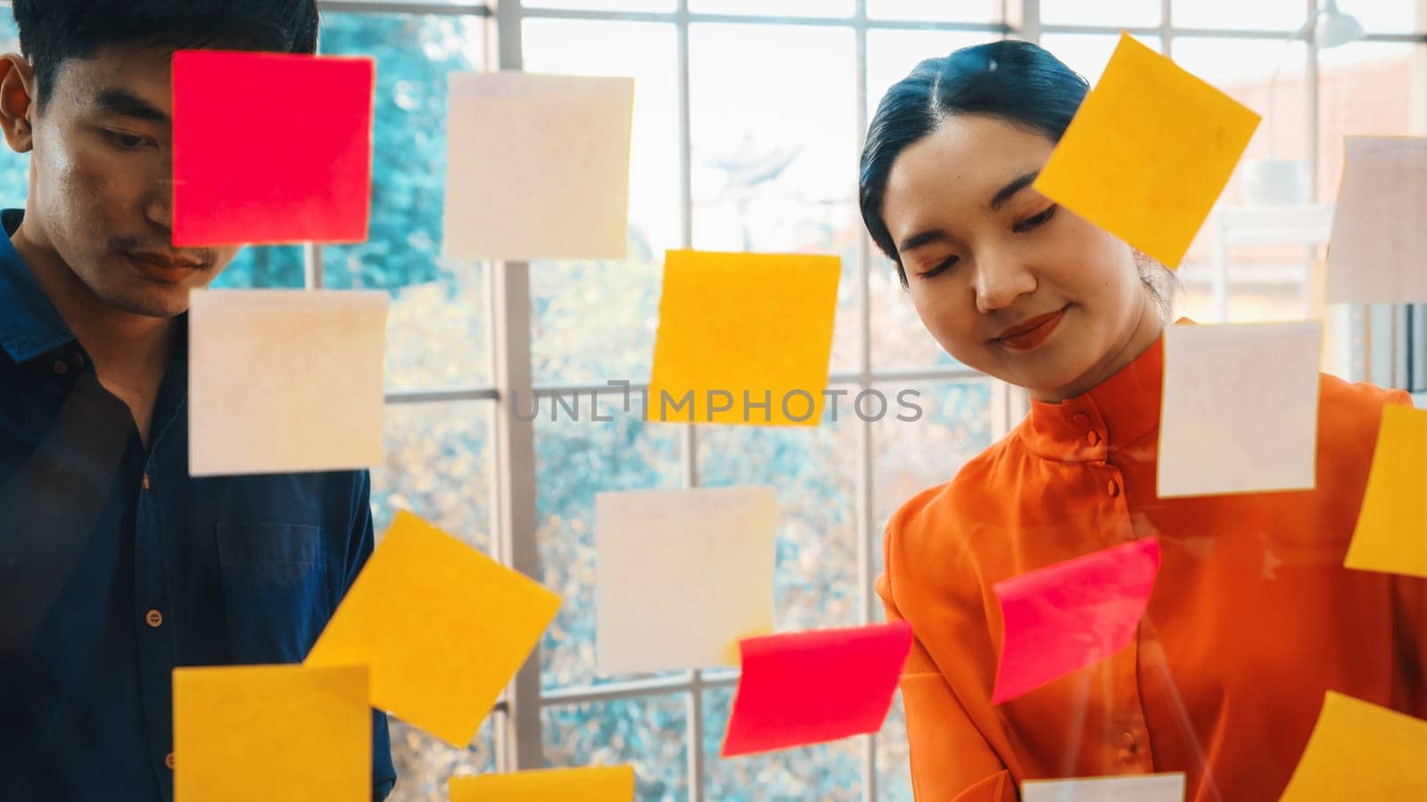 Business people work on project planning board in office and having conversation with coworker friend to analyze project development . They use sticky notes posted on glass to make it organized. Jivy