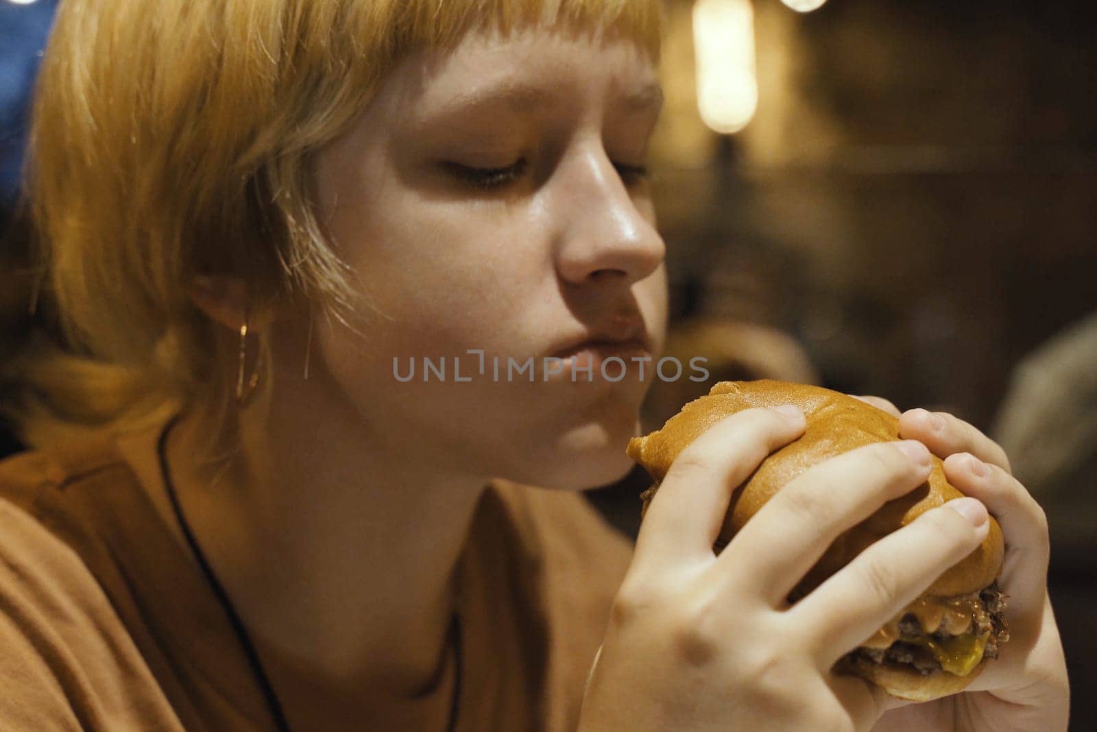 A teenager girl eats a hamburger in a restaurant, enjoying a delicious juicy burger. Close-up.