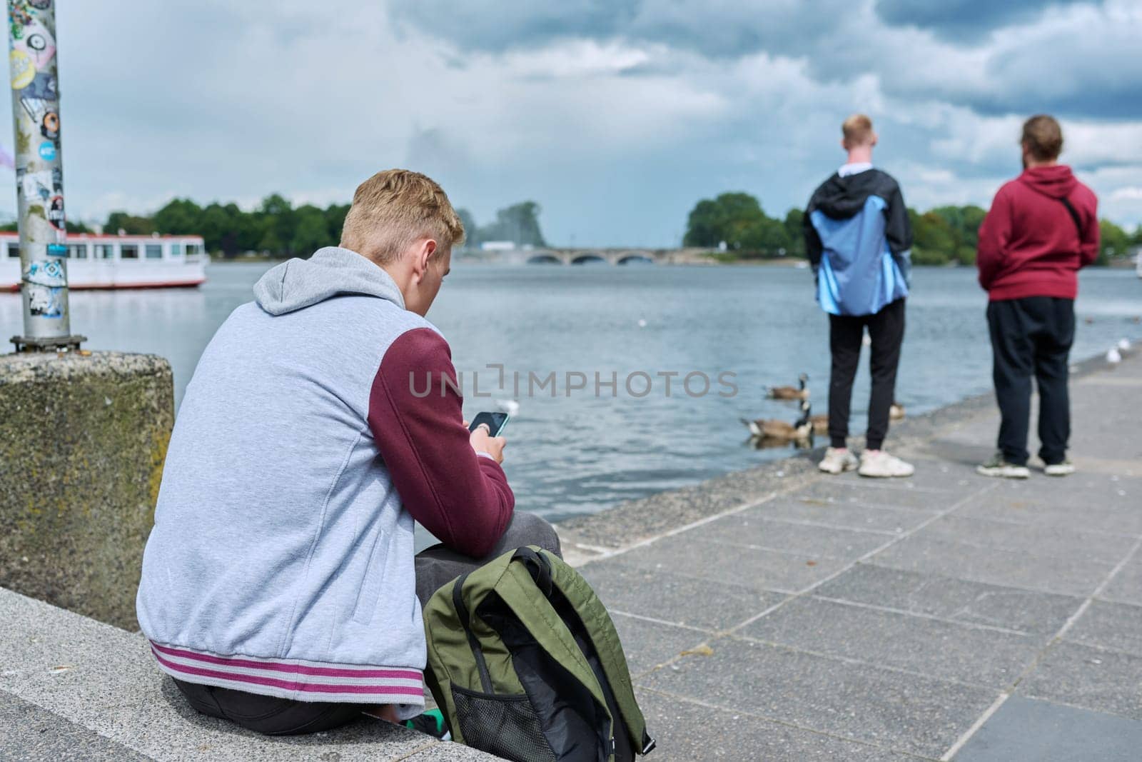 Young guy texting on smartphone, back view, sitting on city embankment by VH-studio