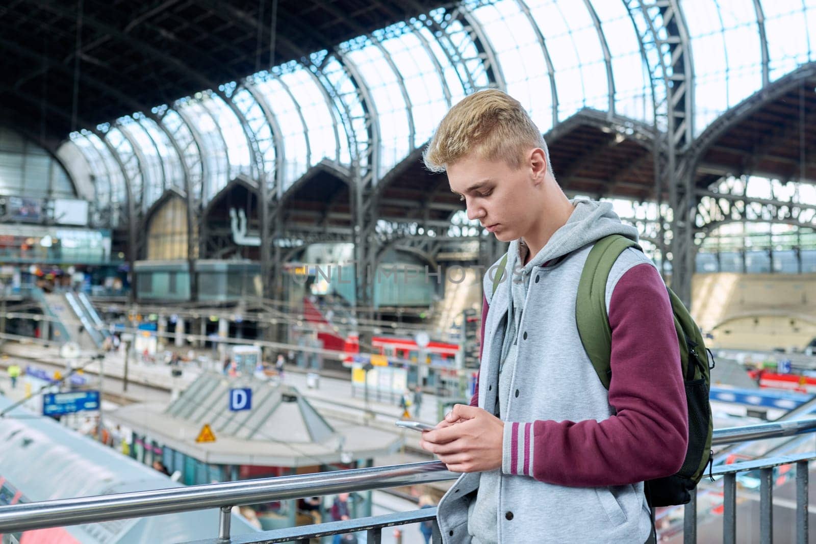 Young serious guy using mobile applications on smartphone standing at railway suburban station. Teenage student looking at train schedule buying electronic ticket. Youth lifestyle, passenger transport