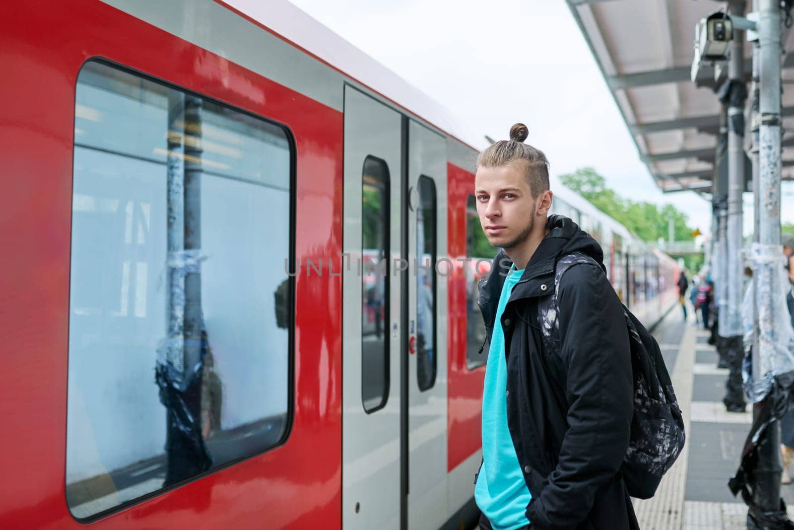 Young male waiting for electric train at city railway platform station. Guy serious student hipster 19, 20 years old with backpack. Urban transport, commuter train, passenger transportation, youth