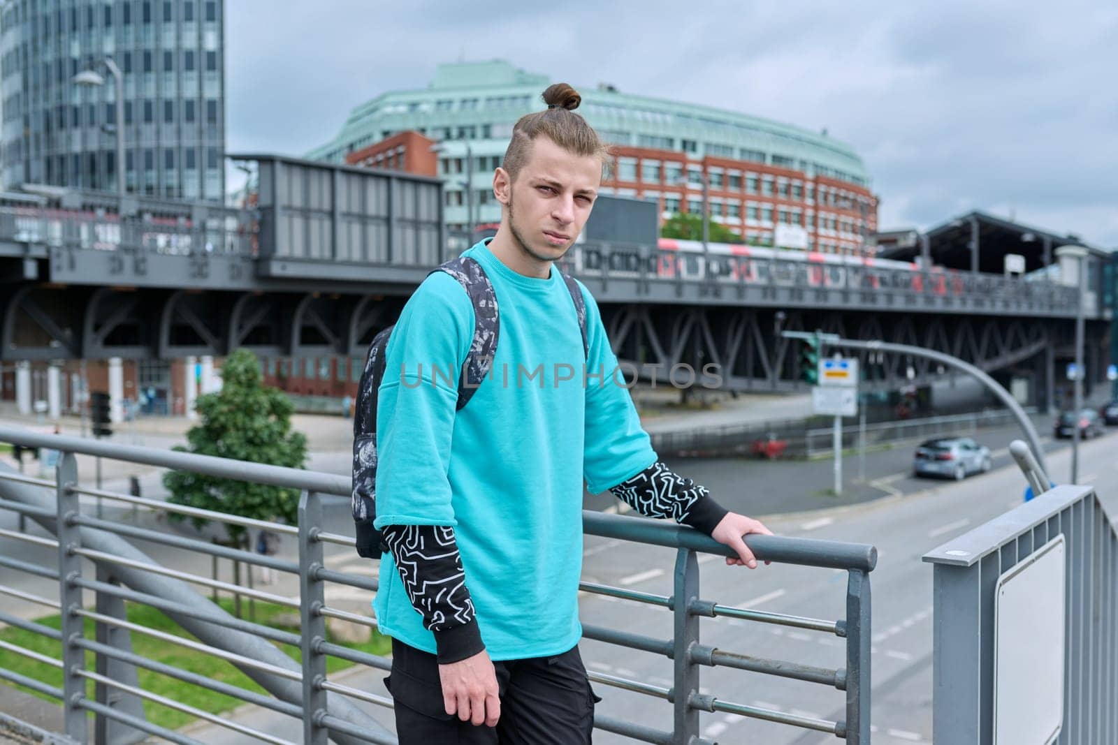 Portrait of handsome guy 19, 20 years old, modern city background. Young male with trendy beard hairstyle, with backpack, urban style. Youth, lifestyle, life concept