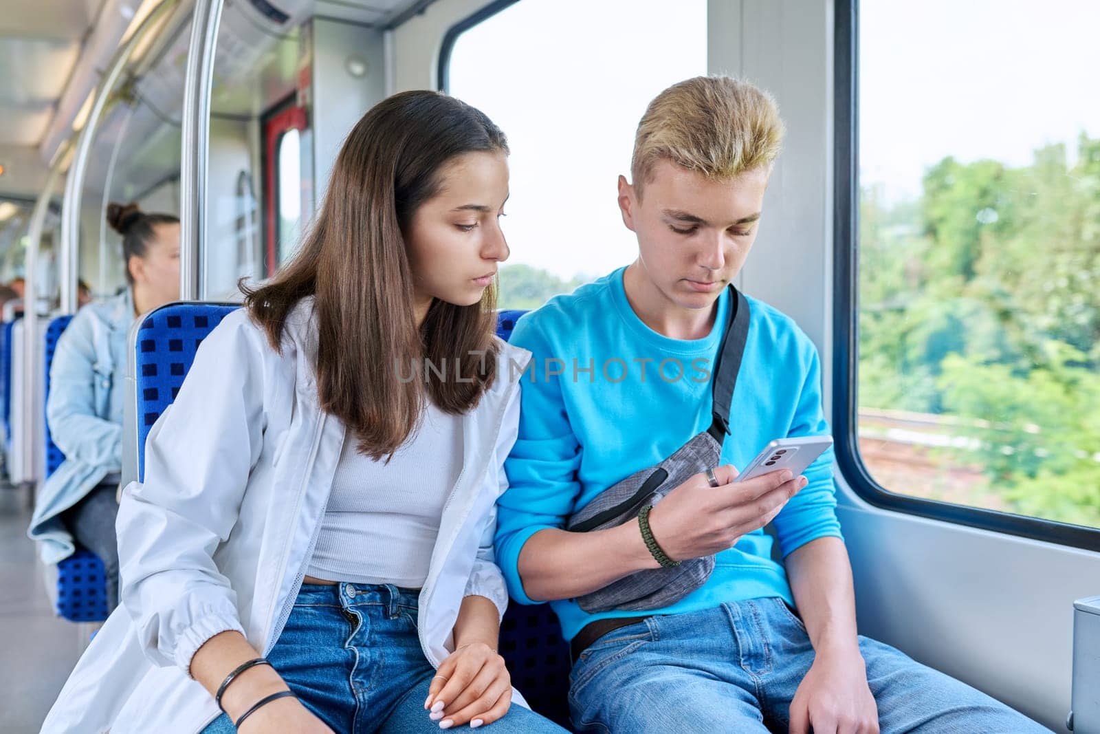 Teenage guy and girl commuter train passengers sitting together looking at smartphone by VH-studio