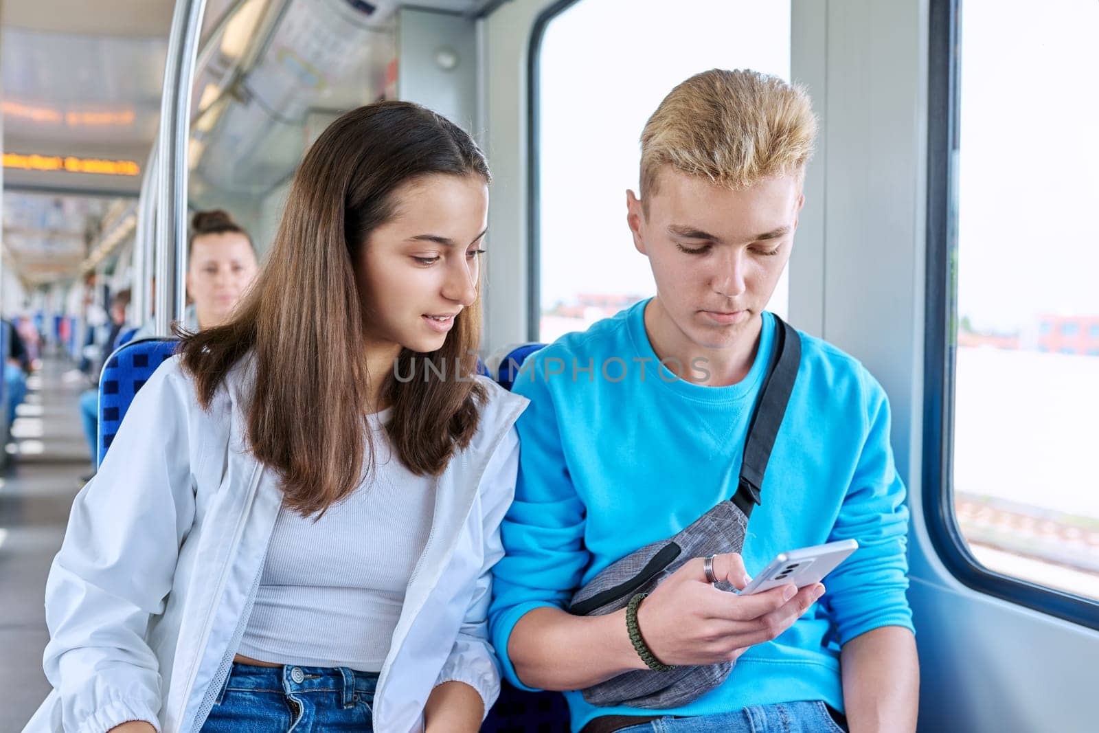 Teenage guy and girl commuter train passengers sitting together looking at smartphone by VH-studio