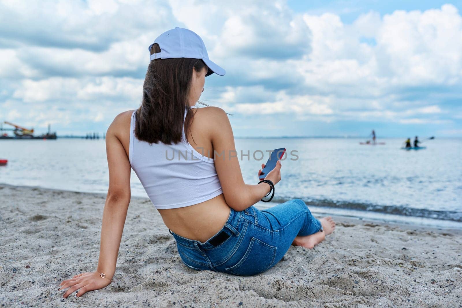 Young female in jeans cap sitting on the beach, using smartphone, back view. Recreation, relaxation, nature, tourism, youth concept