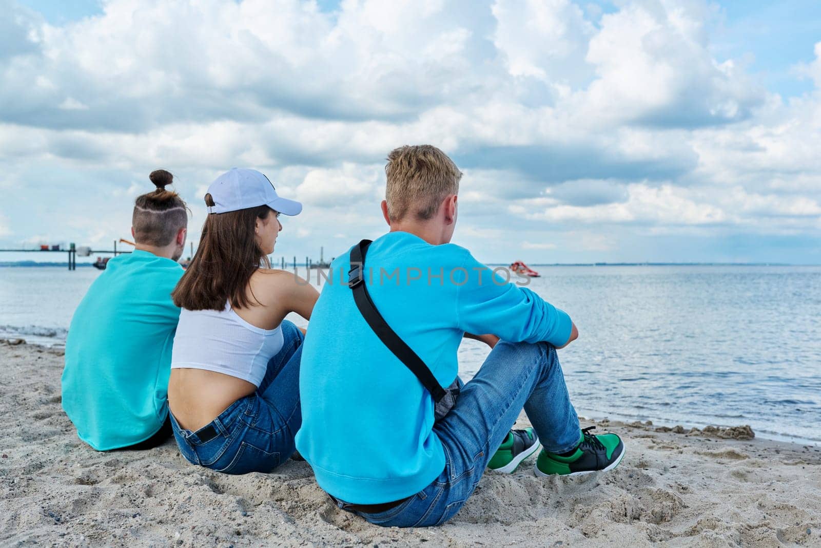 Back view of three teenage friends sitting on the sand at the beach, talking looking at sea. Youth guys and girl, summer vacation leisure friendship fun summertime happiness joy lifestyle holiday
