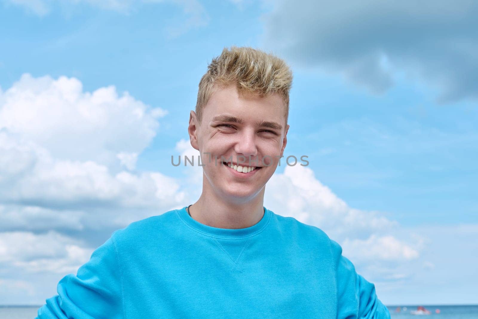 Headshot portrait of handsome smiling teenage guy, face of positive young male against blue sky in clouds. Youth, 18, 19, 20 years of age, lifestyle, enjoy fun, holiday vacation, outdoor