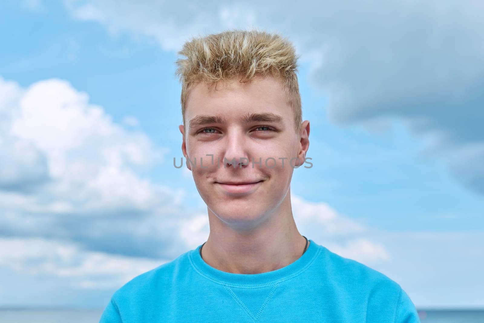 Headshot portrait of handsome smiling teenage guy, face of positive young male against blue sky in clouds. Youth, 18, 19, 20 years of age, lifestyle, enjoy fun, holiday vacation, outdoor