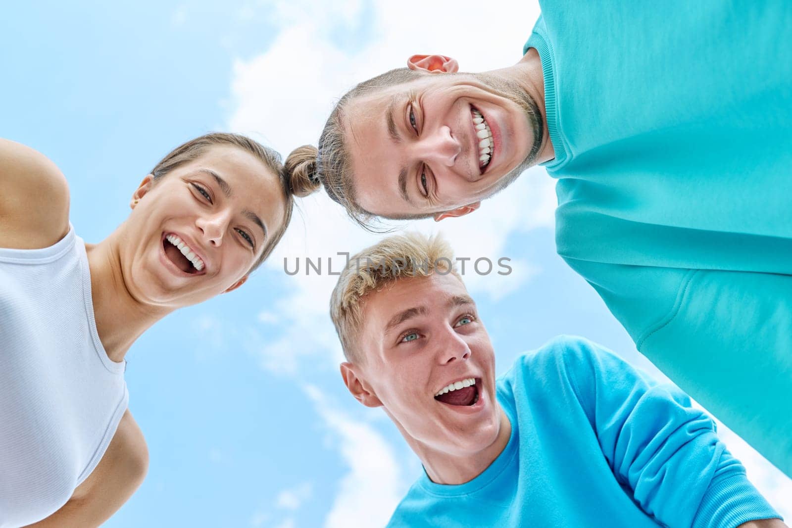 Closeup faces of young teenage guys and girl laughing looking down at camera, blue sky with clouds background. Youth, team summer vacation, communication leisure friendship fun lifestyle holiday