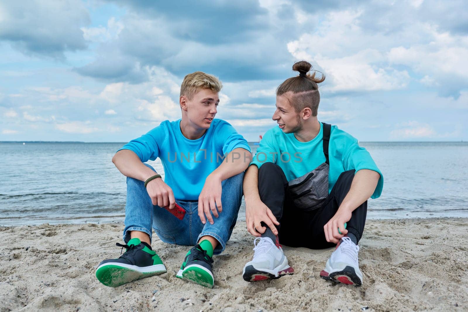 Young teenage male friends sitting on sand at beach, talking, laughing. Youth, summer vacation, communication, relaxing, leisure, friendship, fun lifestyle holiday concept