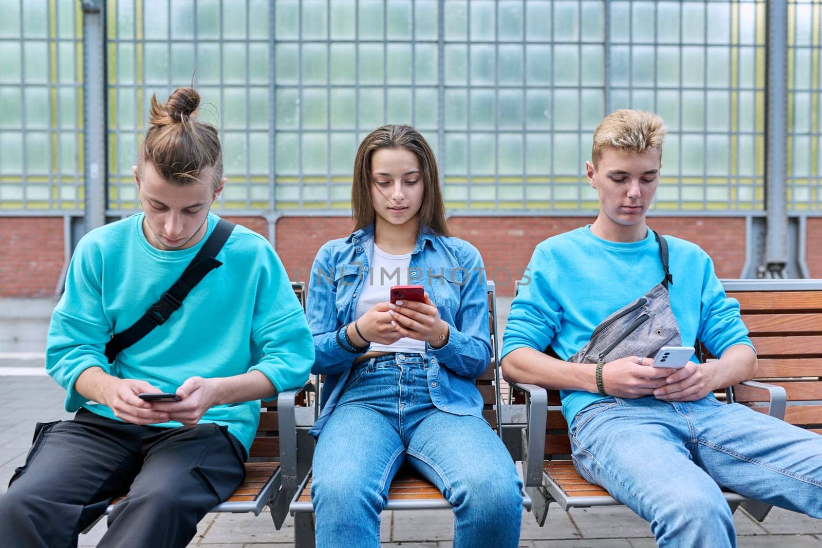 Group of teenage friends using smartphones sitting on chairs on train station platform. Youth, adolescence, digital technology, mobile applications, communication leisure learning social networks