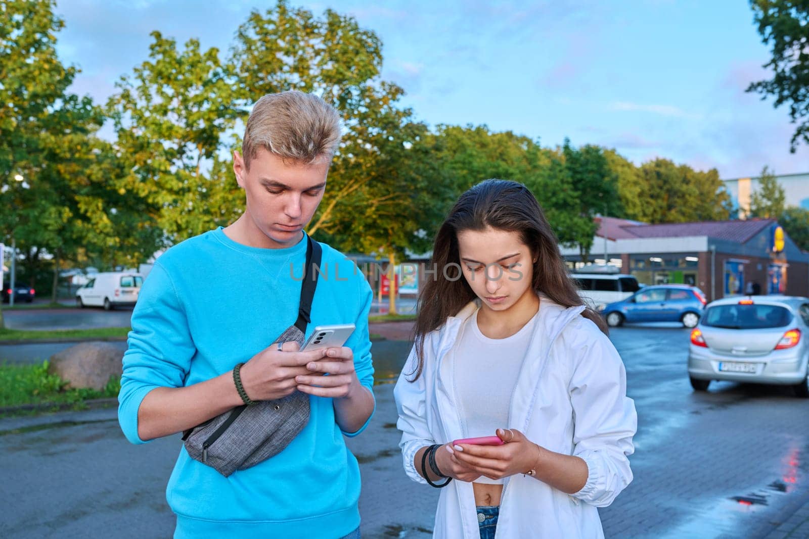 Teenage friends guy and girl standing together holding smartphones, looking at the screen, using mobile phones outdoor. Internet digital technology, mobile applications for leisure study communication