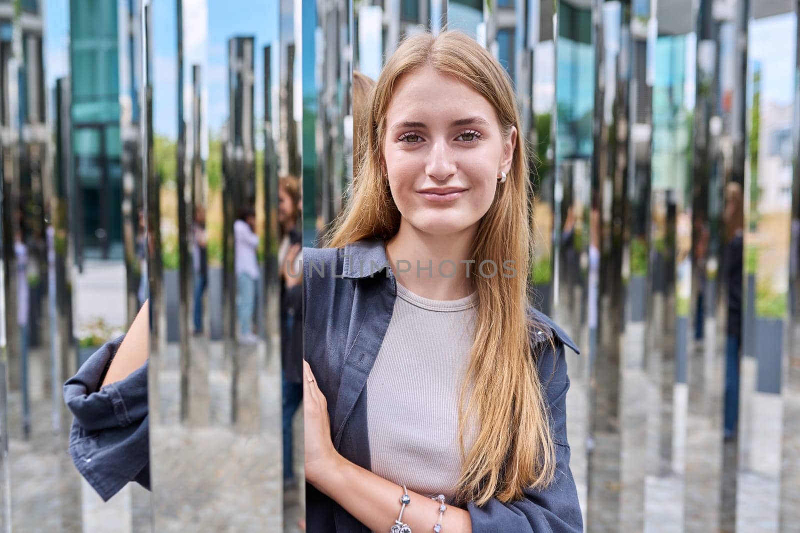 Portrait of young beautiful teenage female in modern city, fashionable smiling girl with long hair looking at camera, on mirror architecture. Beauty, fashion, youth, urban style concept