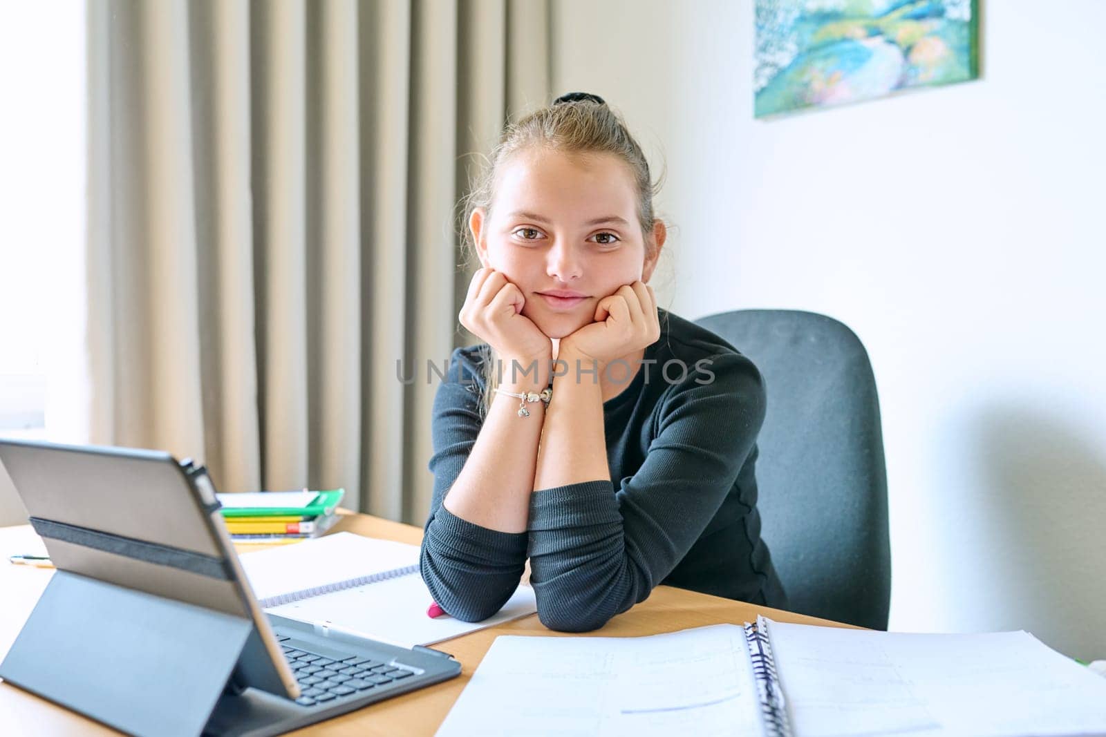 Portrait of smiling child girl sitting at desk at home with digital tablet textbooks, looking at camera. Education, knowledge, school, childhood concept
