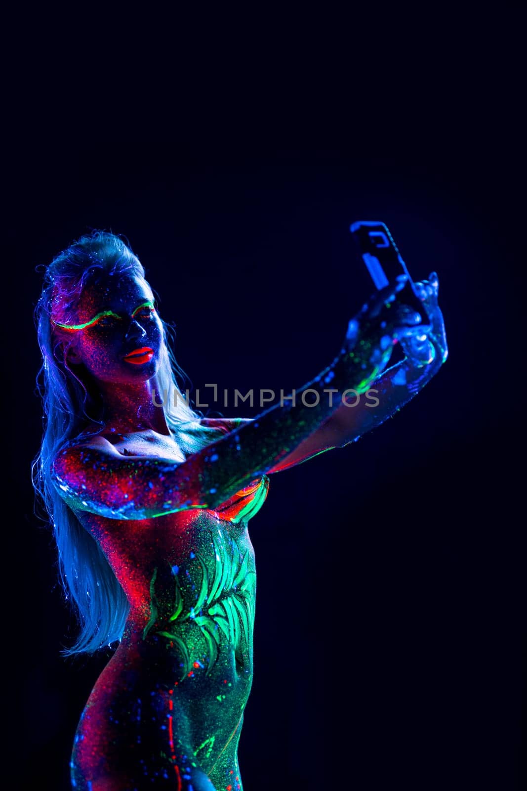 Nude woman with fluorescent colorful body art doing selfie in dark