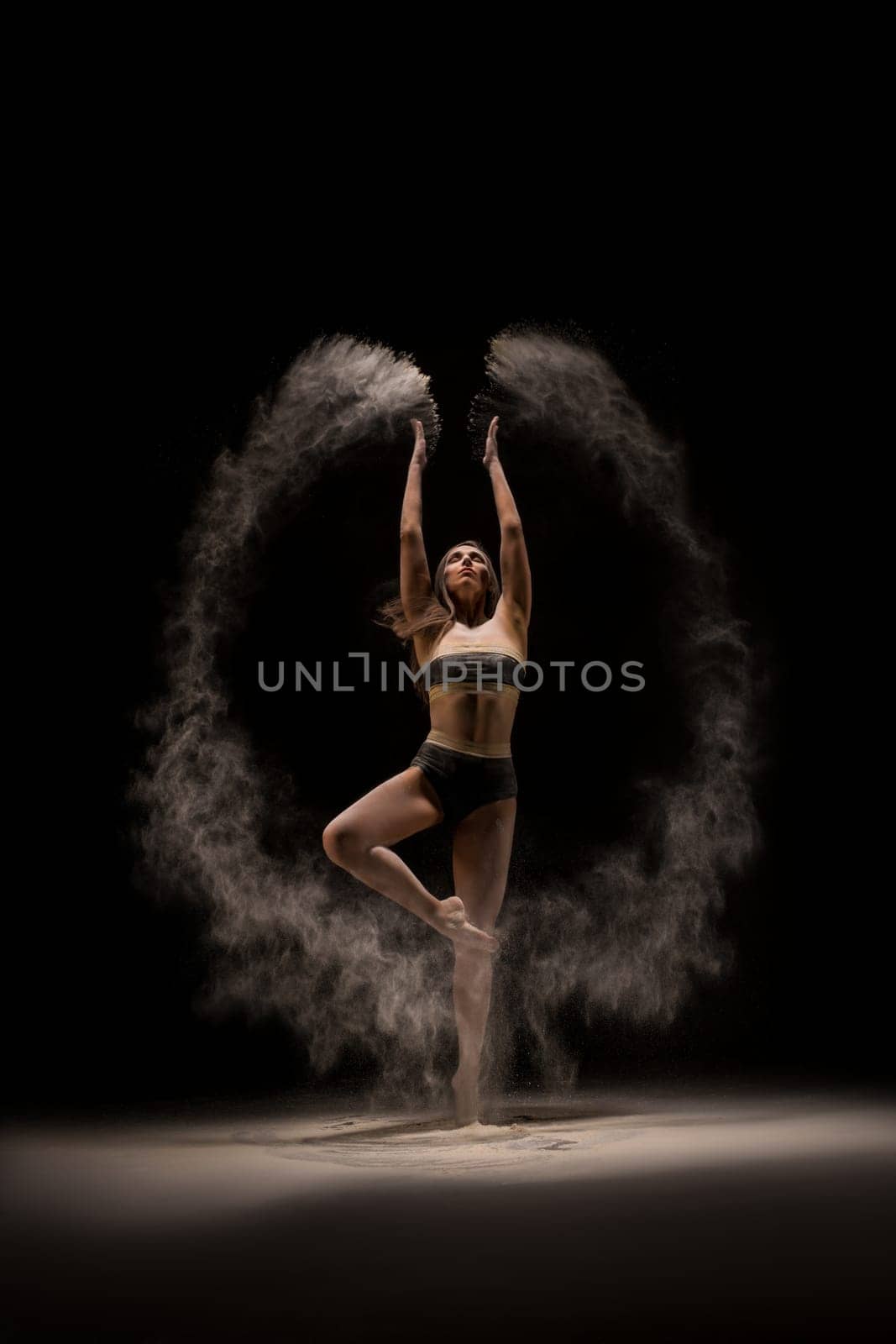 Full length slim female in bra and panties throwing sand in air and dancing against black background