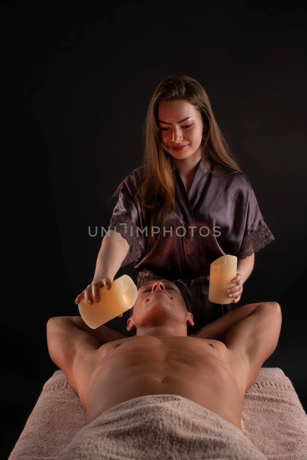 Professional masseuse applying melted wax from candles on chest of relaxed male client during spa procedure in salon