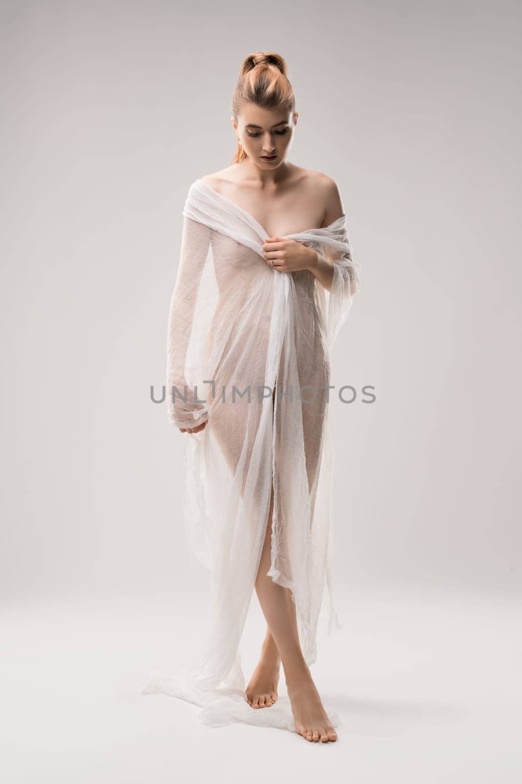 Graceful nude female covering body with transparent cloth standing on white background in studio and looking down