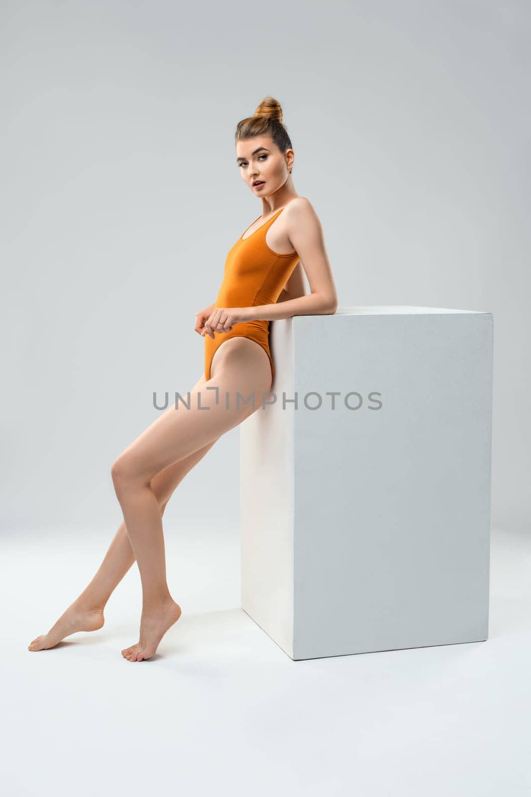 Full body side view of slim barefoot young female gymnast in orange leotard leaning back against white block while standing in light studio