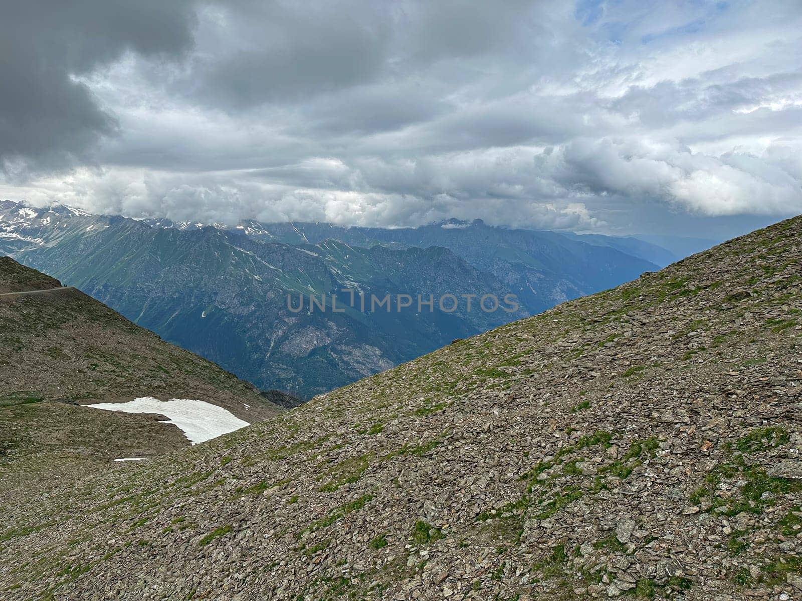 A vast mountain view with rocky terrain and patches of snow. Dark clouds loom overhead, framing the peaks and valleys of a remote alpine region, suggesting a change in weather.