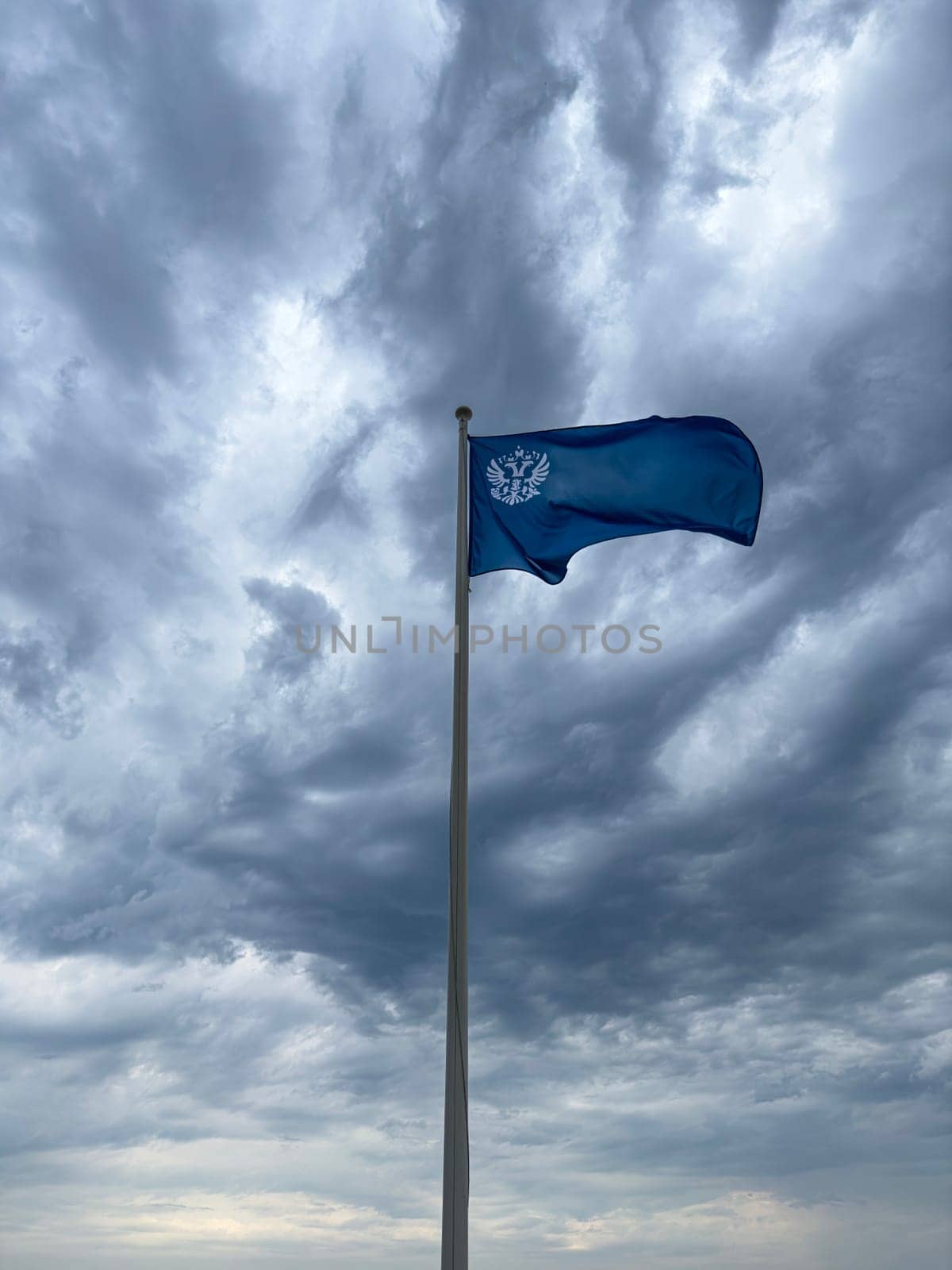 A vibrant blue flag flutters atop a tall pole under a swirling gray sky filled with clouds. The contrast between the flag and the turbulent weather creates a striking visual.