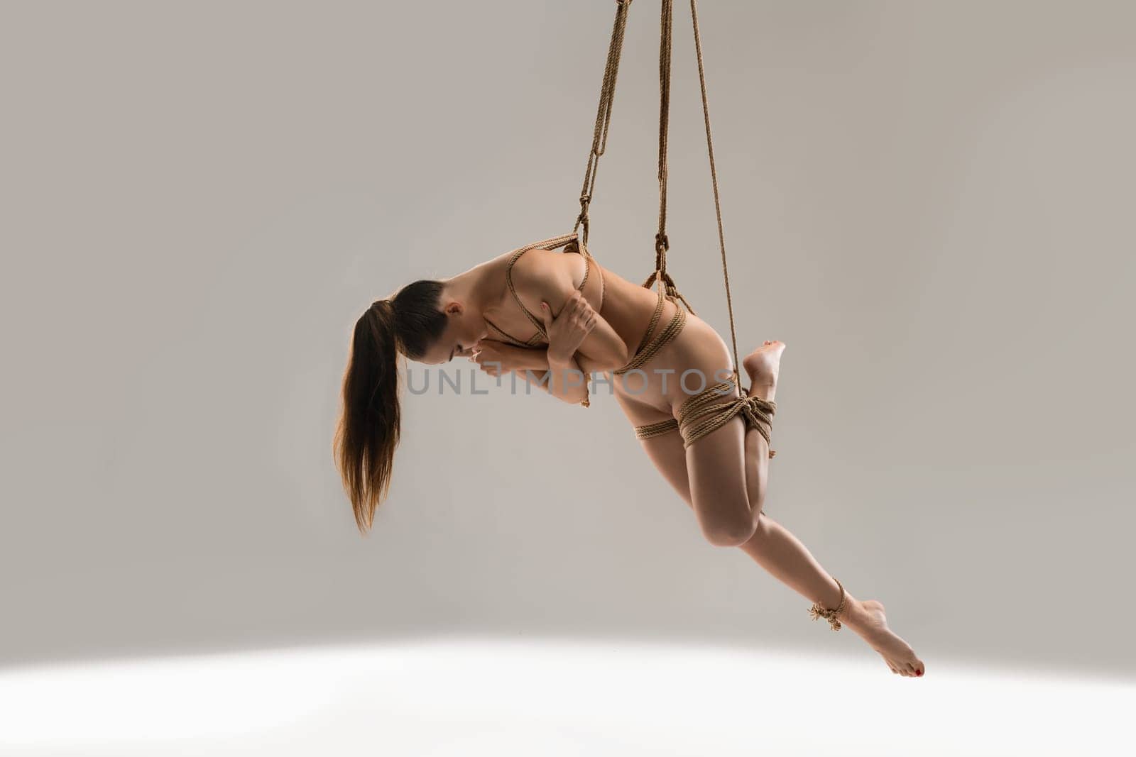 Full body nude barefoot woman with ponytail hanging on Shibari ropes while practicing BDSM against gray background