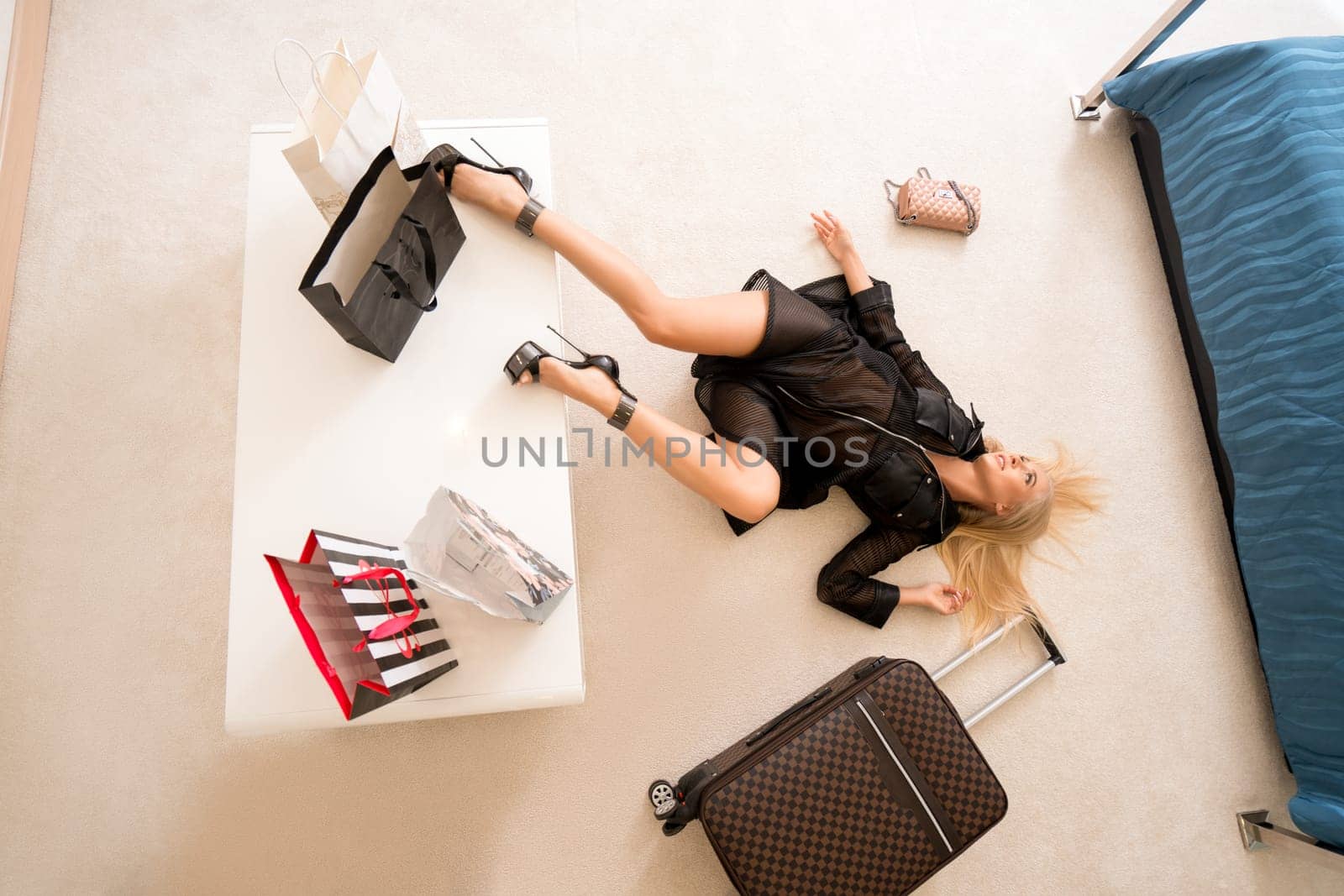 Top view of sensual slim blonde female in black lingerie and high heels lying on floor near suitcase in light hotel room