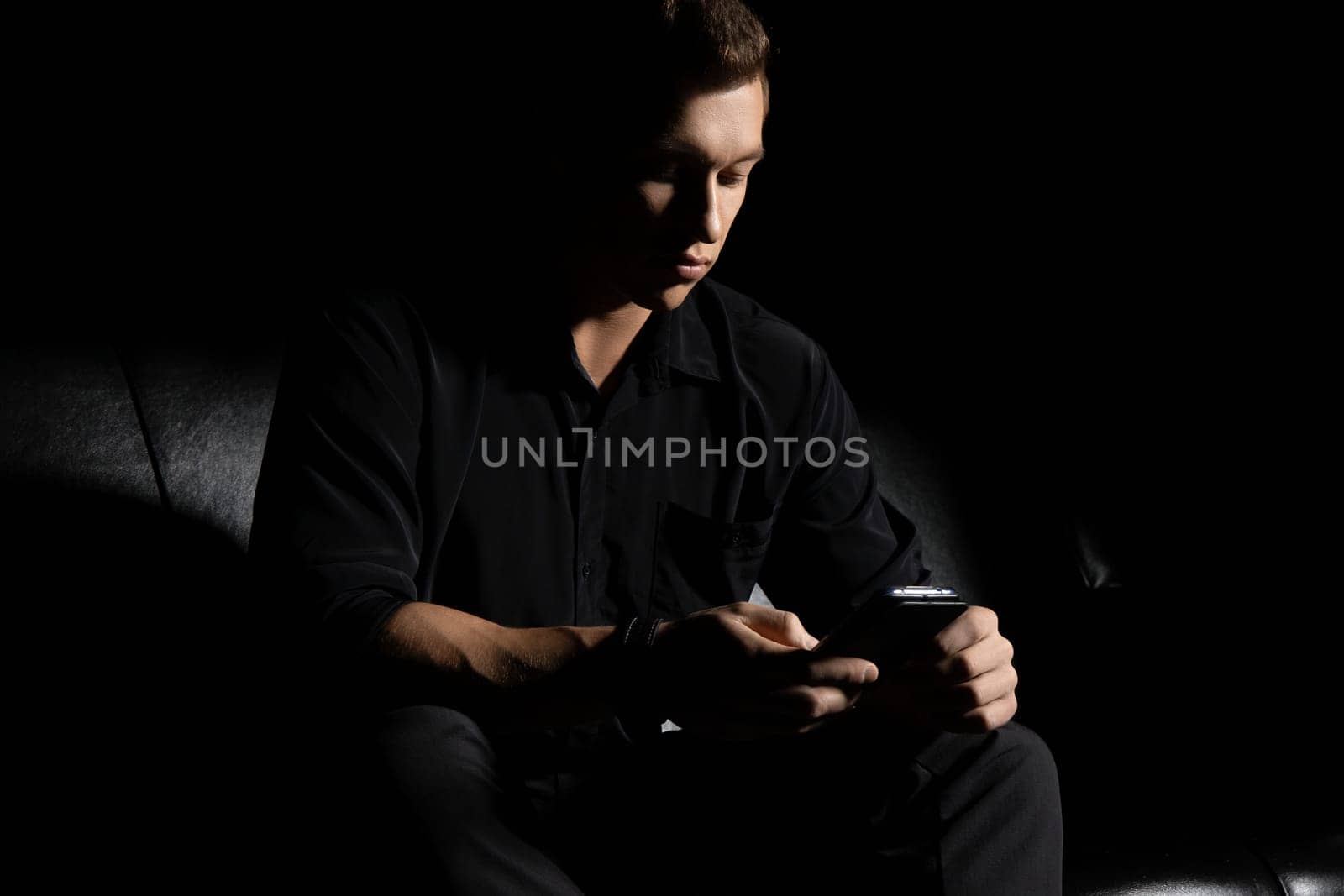 Concentrated young man in black outfit sitting on leather couch and browsing mobile phone in dark studio