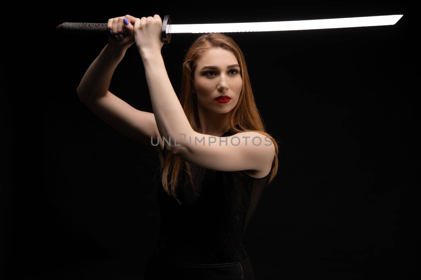 Determined young female in black outfit with red lips holding katana sword in raised hands while standing against black background