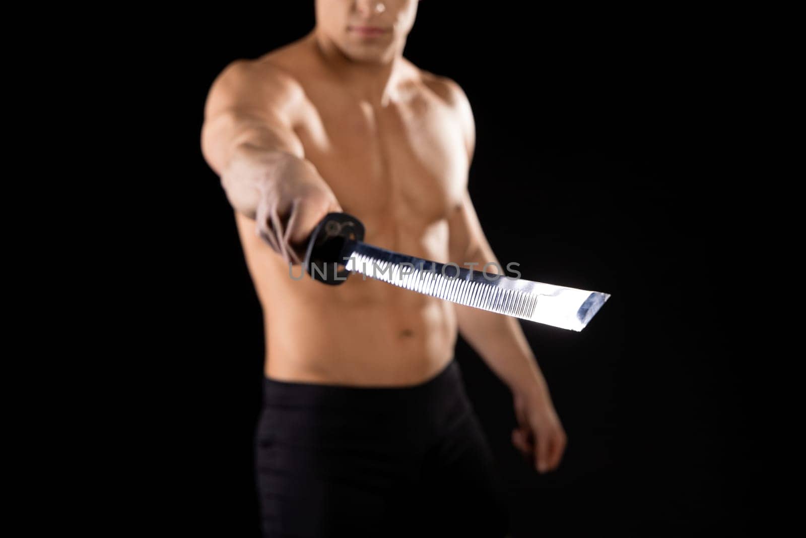Crop unrecognizable muscular male with naked torso holding Japanese katana sword in outstretched arm against black background