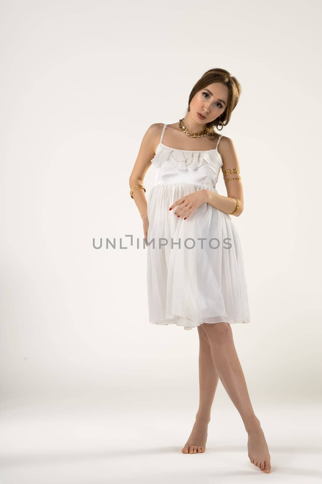 Full body charming barefooted young female model in elegant white dress and jewelry looking at camera while siting in white studio