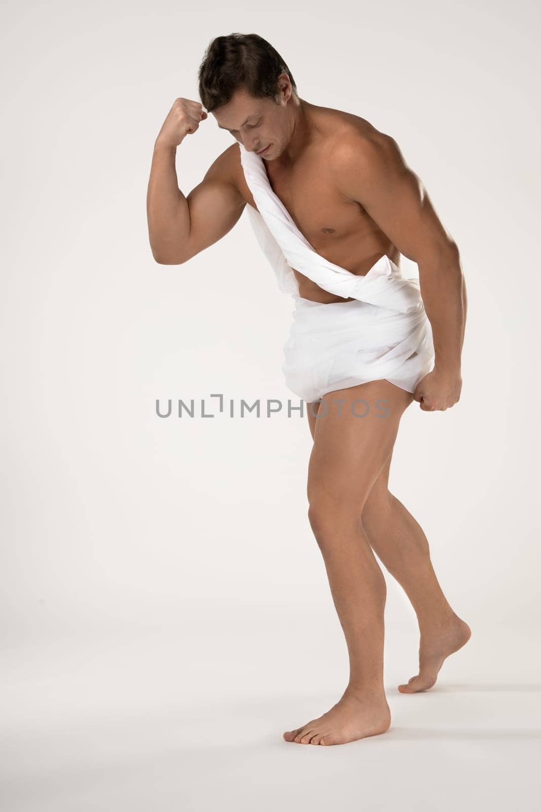 Full body of young muscular bare chested male wrapped in white cloth in antique style standing in white studio