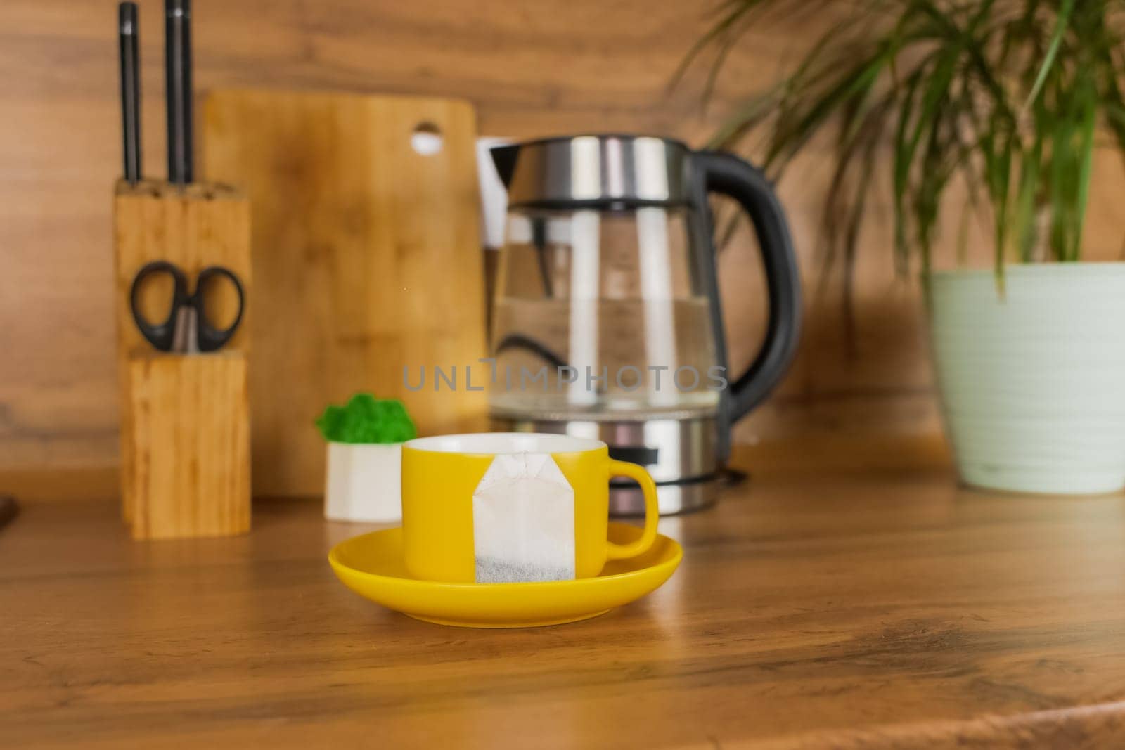 A yellow cup of tea stands on a yellow saucer and on the saucer lies a tea bag on a wooden table against the background of a teapot, a plant and kitchen utensils. The concept of tea drinking, break. High quality photo