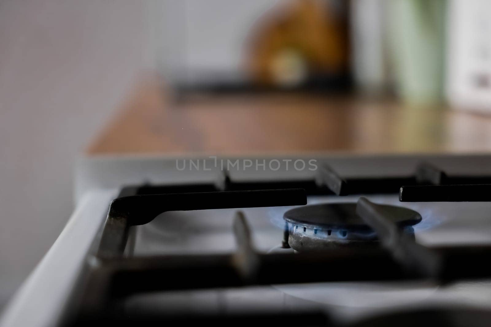 Close-up of a white gas stove with a black cast-iron grate and a black burner on which a blue fire burns against the background of a wooden kitchen in out-of-focus. Gas stove, cooking concept. High quality photo
