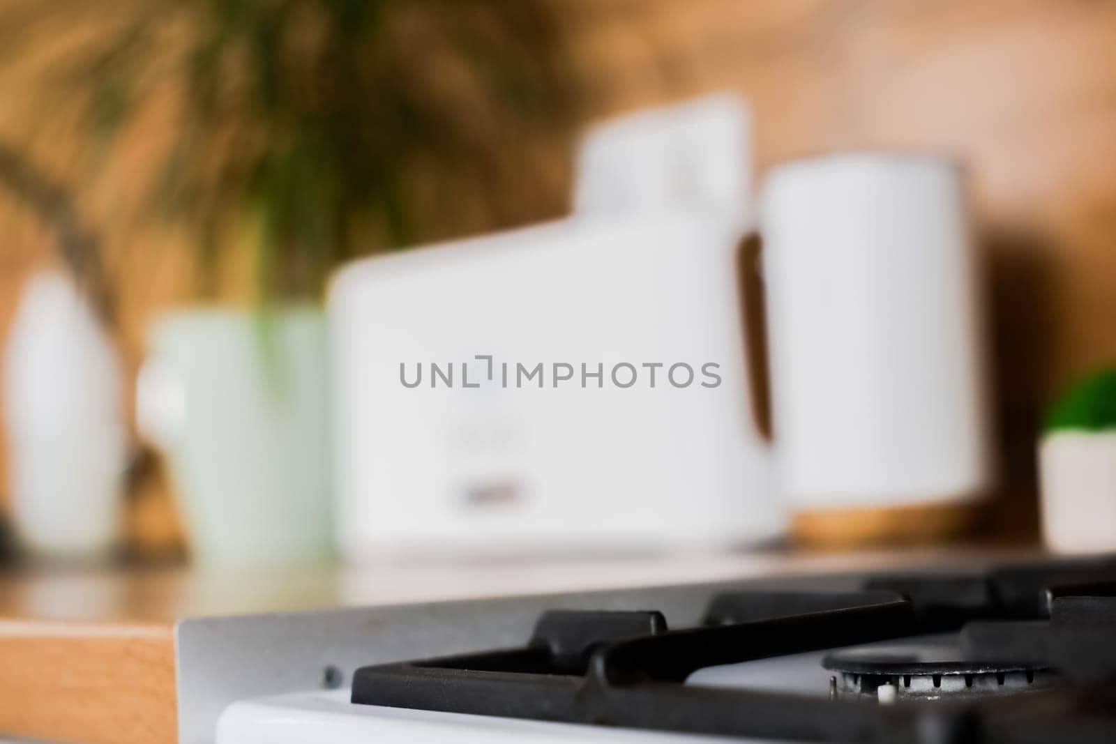 Close-up of a white gas stove with a black cast iron grate and a black burner on which a blue fire burns against the background of a wooden kitchen in out-of-focus with kitchen utensils and a plant. Gas stove, cooking concept. High quality photo
