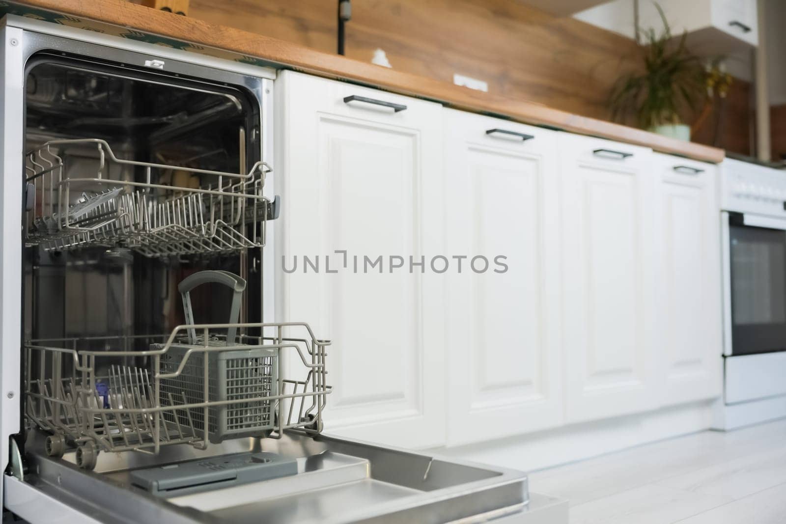 An empty open dishwasher with gray drawers for dishes and cutlery stands on the left of the photo against the background of a white wooden kitchen in a slight out of focus. Dishwashing concept, comfort. High quality photo