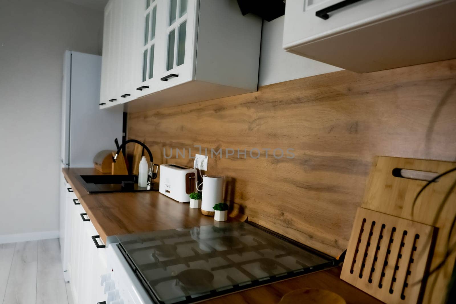 Side view of the modern kitchen, wooden countertop with kitchen appliances and appliances, white cabinets, gas hob, white fridge, black sink and plants. High quality photo