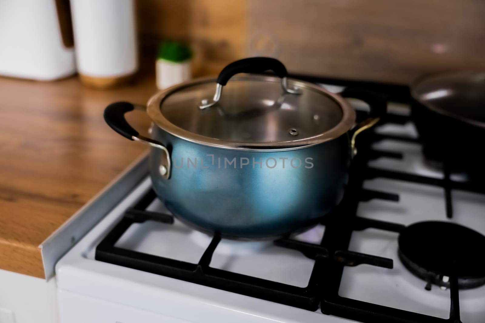 On a white gas stove with a black cast iron grate there is a blue pot with a glass lid and rubber pads on the handles against the background of the kitchen in out-of-focus, Concept of cooking, cooking, cooking. High quality photo