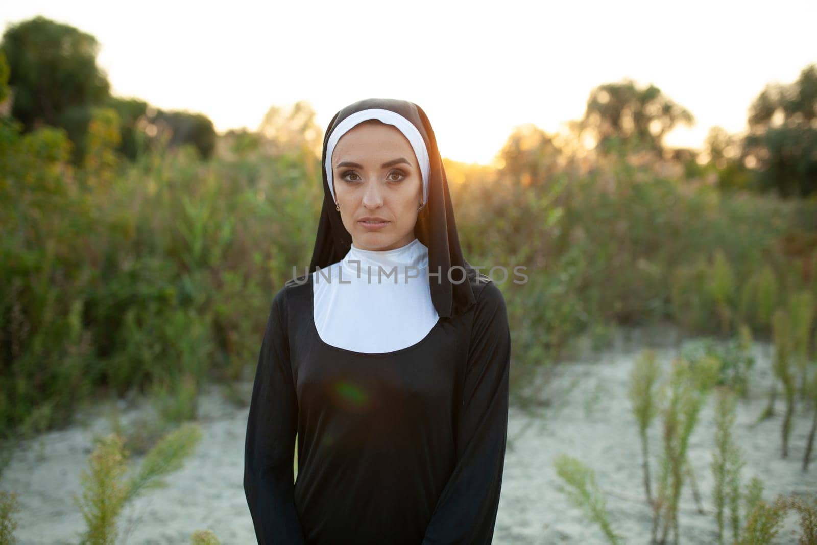 Serious nun looking at camera while standing on field during sunset. Young woman in religious clothes is surrounded by plants outdoors