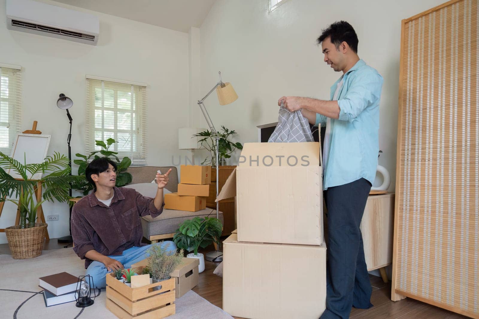 A gay couple in their new home, one unpacking boxes while the other points out decor ideas, showcasing teamwork and creativity.