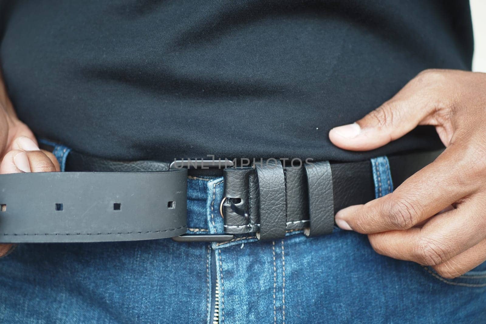 A man adjusts his black leather belt while wearing blue jeans.