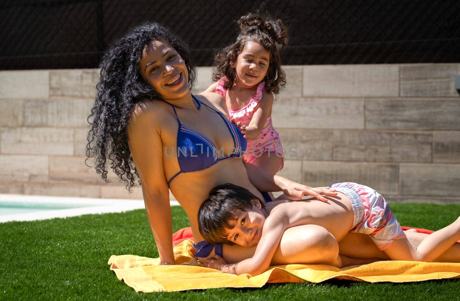 Happy mother in blue bikini relaxing by the pool with her two children on a sunny day