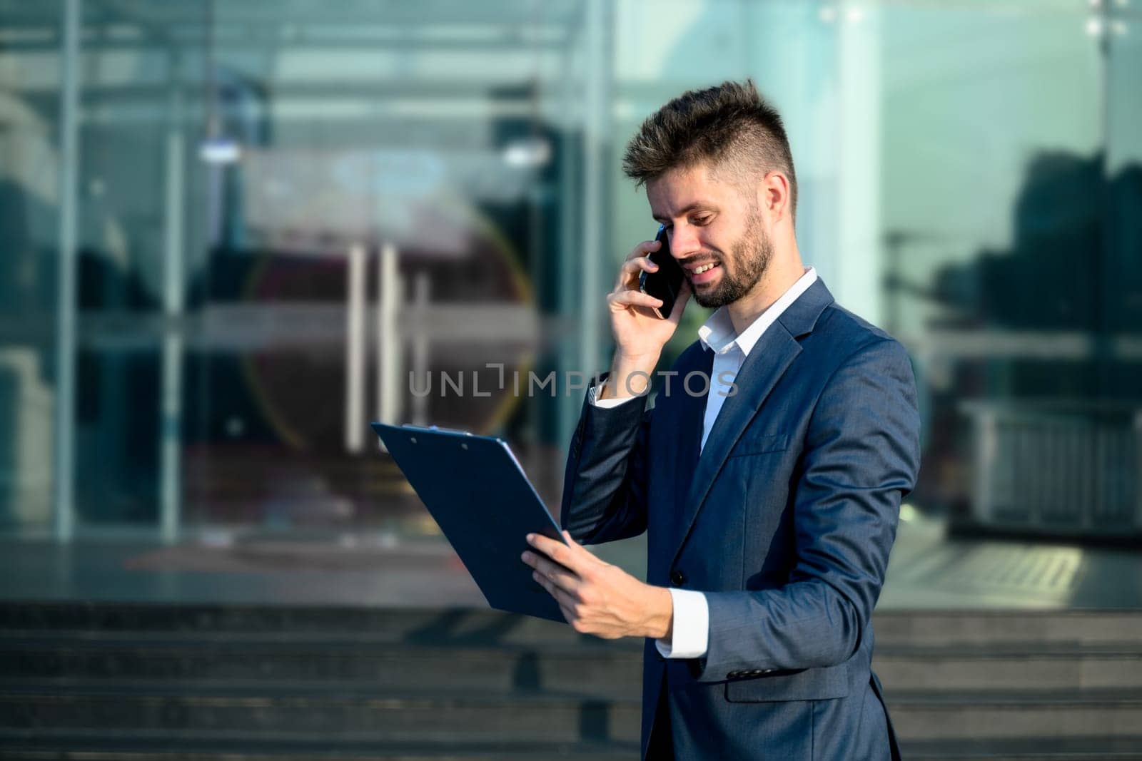 Businessman holding a clipboard and talking on smartphone outside modern office building.