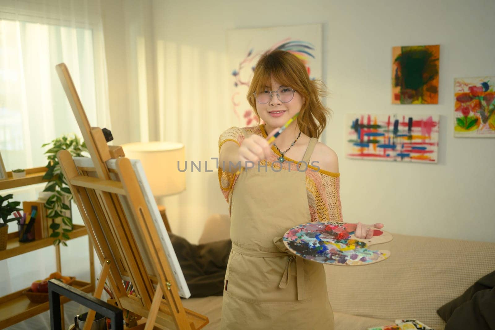 Cheerful female artist holding a palette and extending paintbrush towards the camera.