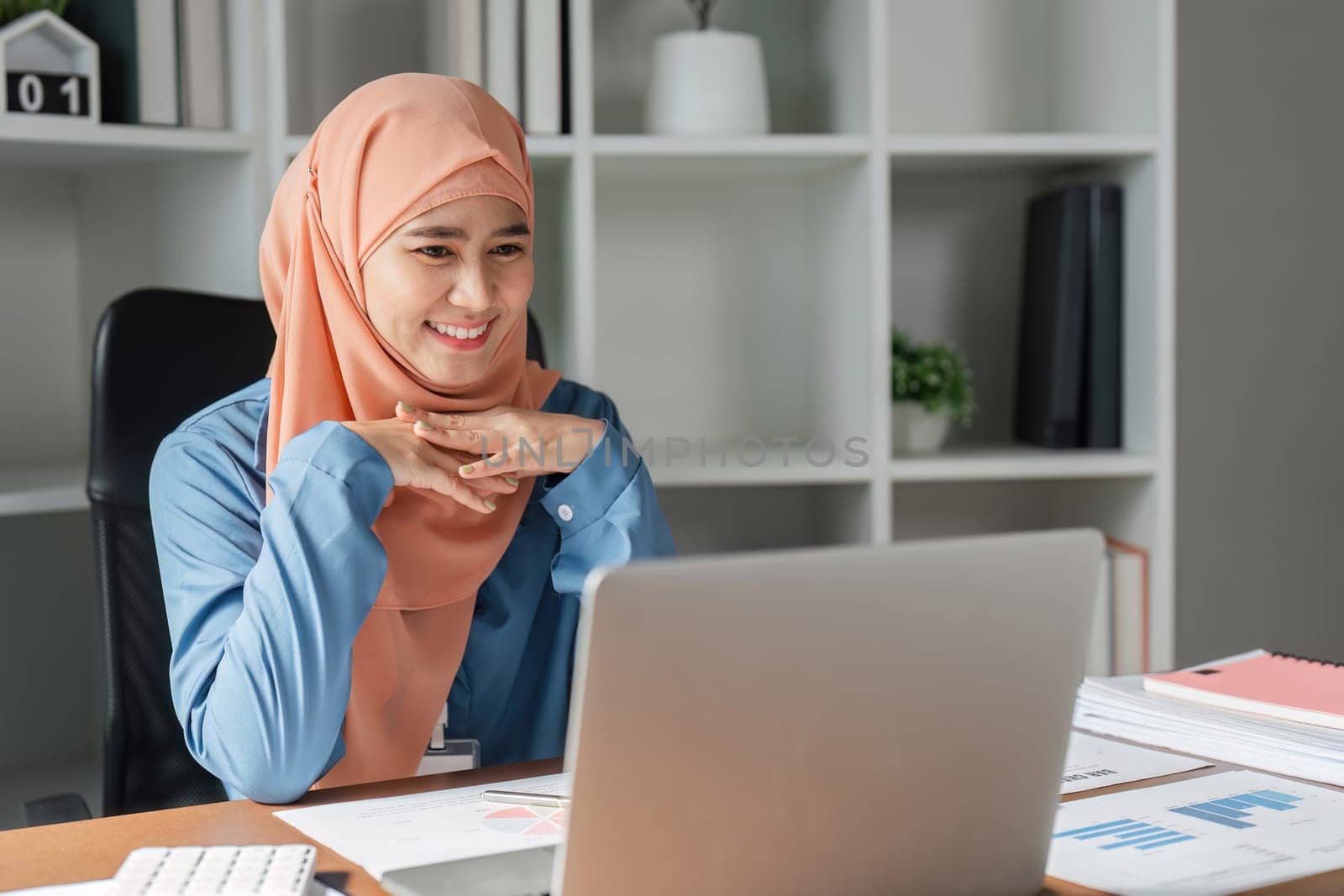 A Muslim woman in a hijab, happily engaging with her laptop and reviewing documents in a contemporary office workspace.