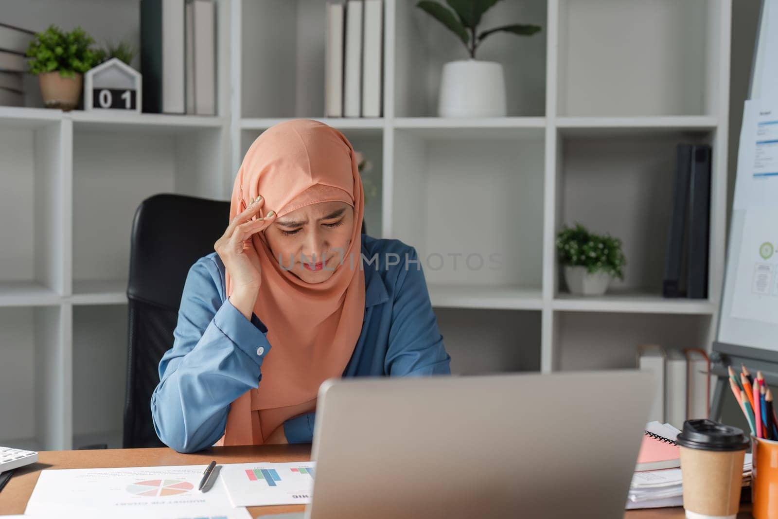 A diverse Muslim woman in a hijab appearing overwhelmed and stressed while focusing on her laptop in a modern office filled with financial documents.