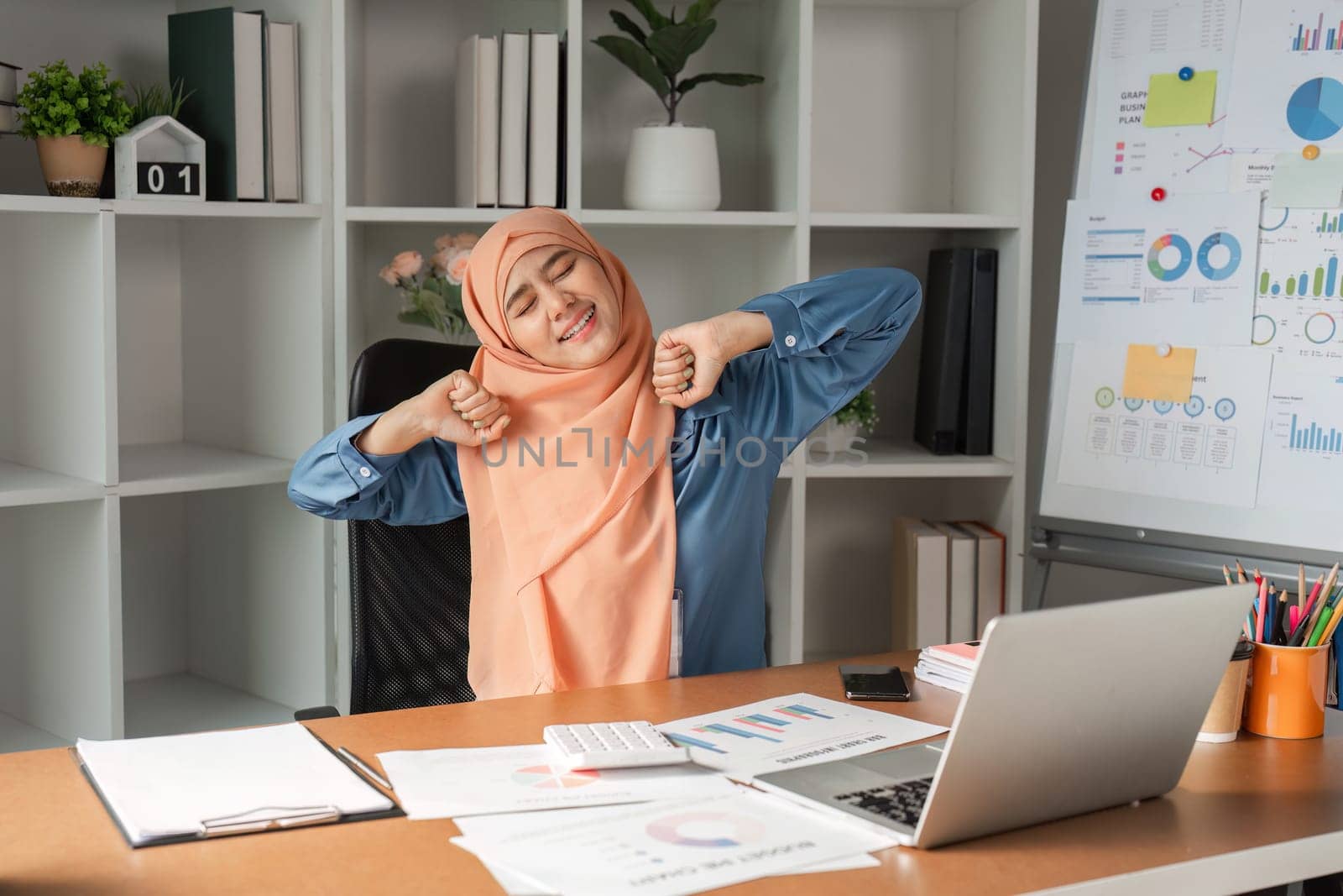 A Muslim woman in a hijab stretching at her desk, taking a break from managing business reports and financial documents in a professional setting.